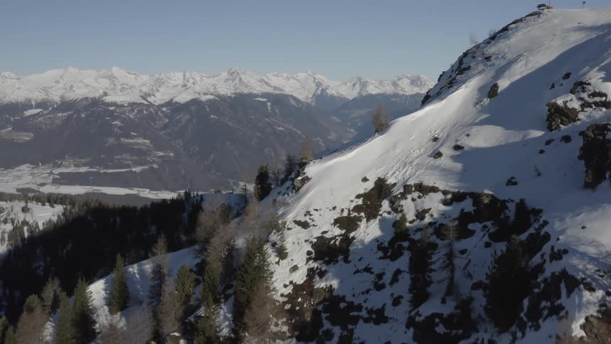 Snow covered forested mountain and valley with a snow covered mountain range in the distance under a clear sky