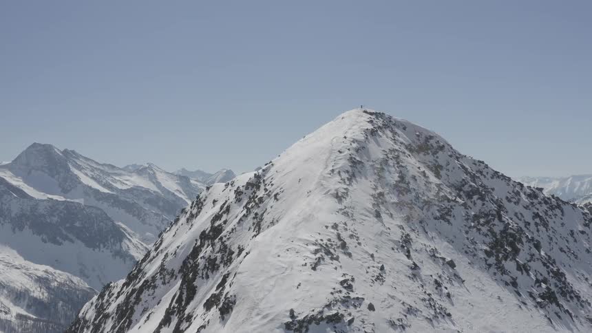 A person is standing on the peak of a snow covered mountain under a clear blue sky