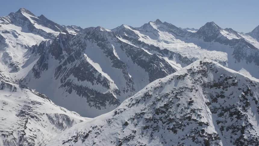 Two people are standing on the peak of a snow covered mountain near a snow covered mountain ridge under a clear sky