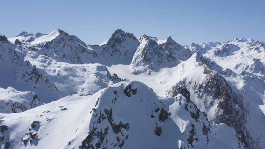 A group of people stand atop a snowy mountain peak high in the mountains on a clear day
