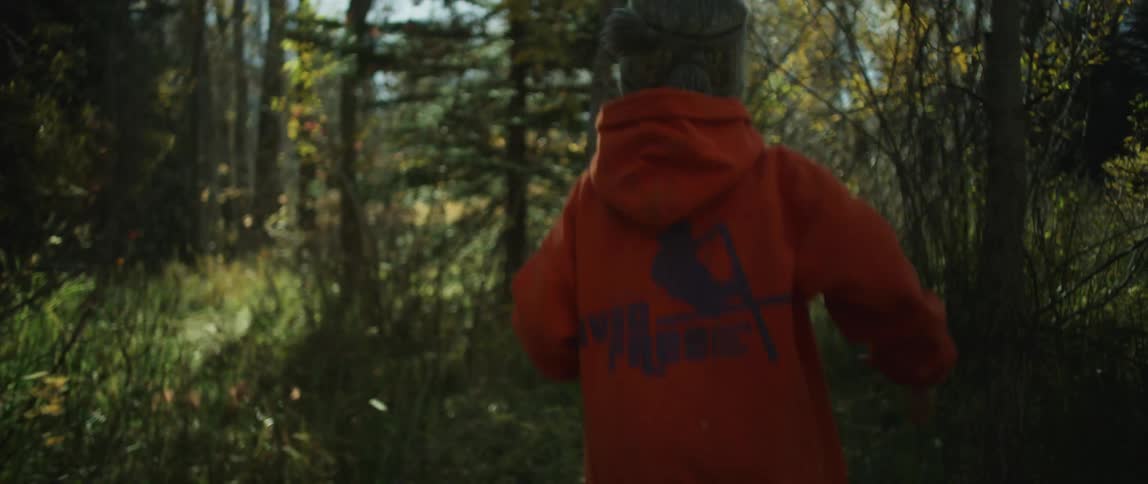 A child playfully runs through a woodland while swinging a stick at trees