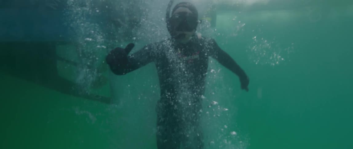A man with snorkeling equipment on jumps off a boat in to the water and goes under water and smiles as a woman sits on the boat putting flippers on