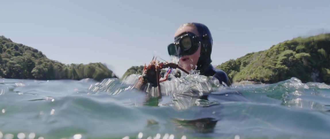 Young boy wearing a wet suit rises to the surface of the ocean with a lobster on his hand