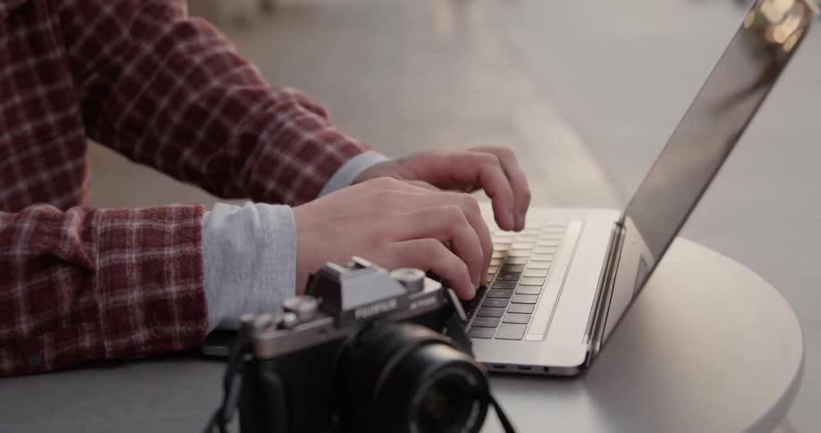 A person sitting at a small table types on their laptop with their camera positioned nearby