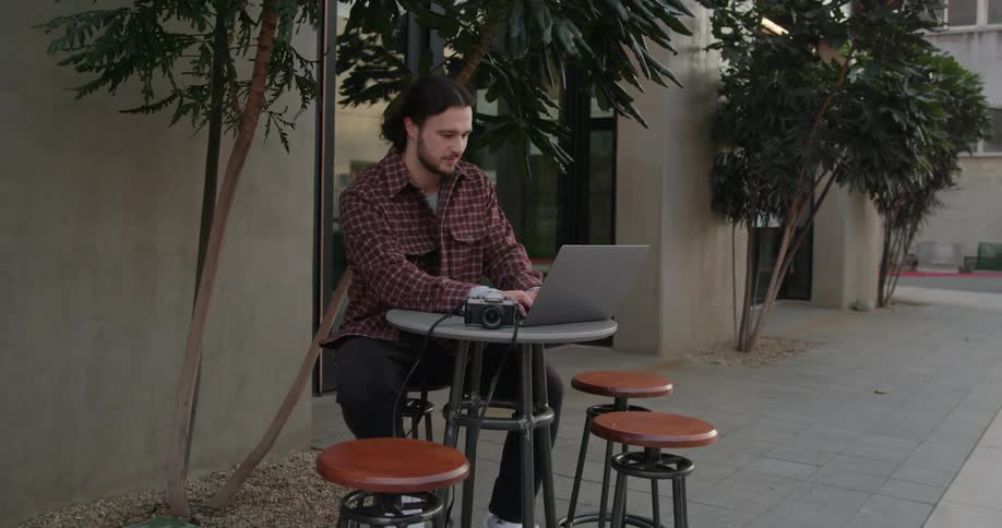 Photographer sits at outdoor table on sidewalk with laptop as car passes in background