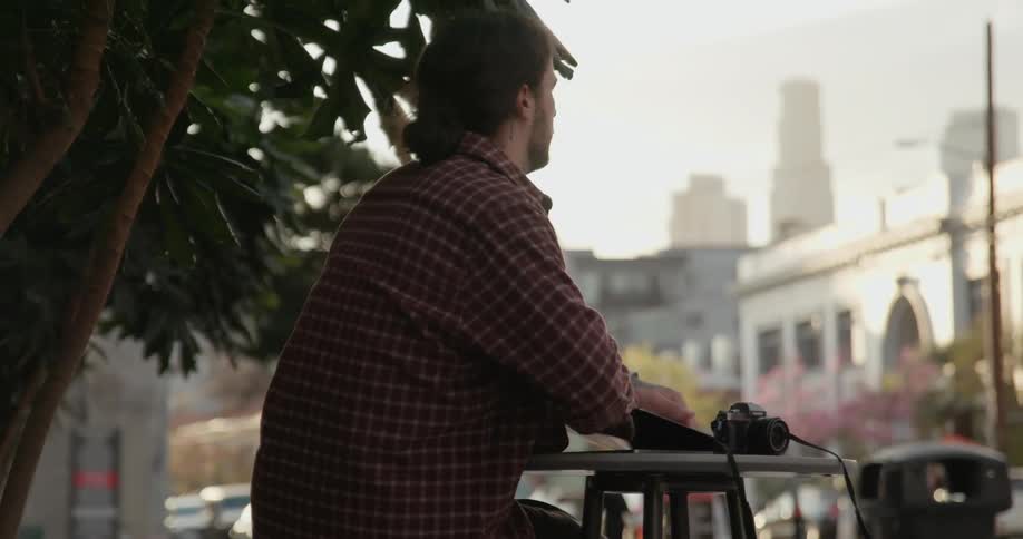 Man carrying camera and laptop sits at outdoor table as another pedestrian walks onto street in the background