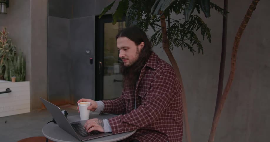 Photographer sits at outdoor table drinking from cup and using laptop as pedestrians approach along the sidewalk