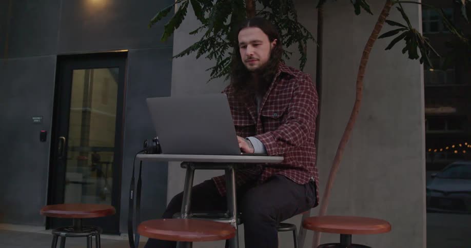 Young long haired man sitting at a table on the city street drinks a beverage while working on a laptop