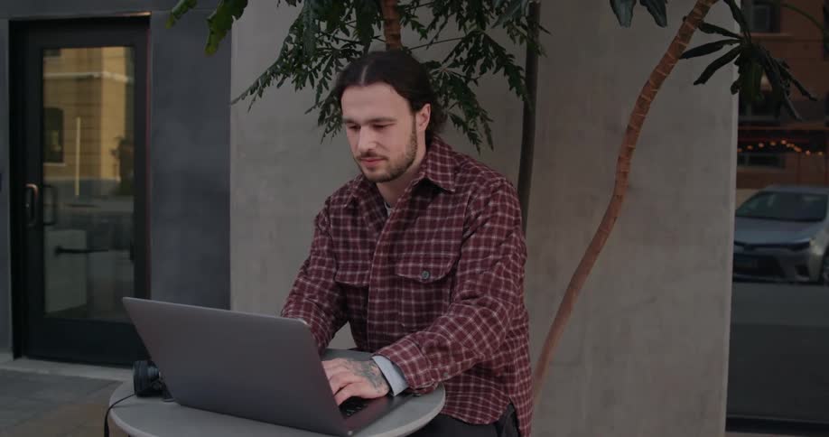 Young long haired man sitting at a table on the city street drinks a beverage while working on a laptop
