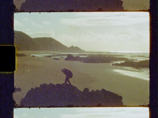 A person climbing the jagged edge of a rock promontory near the beach at sunset caught on vintage film