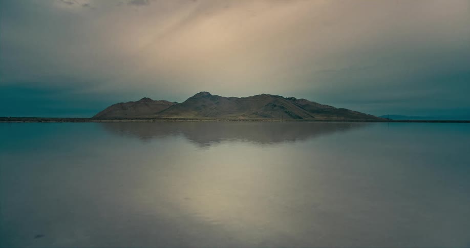 A mountain sits behind a rippling body of water on a cloudy evening