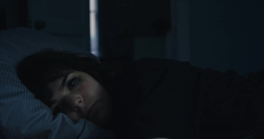 A woman with mascara running down her face lies in bed with the curtains drawn