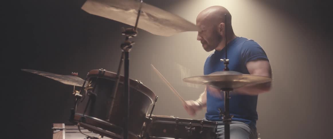 A man plays the drums with a light shining behind him in a dark space