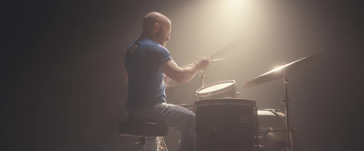 Bald male drummer with blue shirt jeans and beard plays drums in spotlight