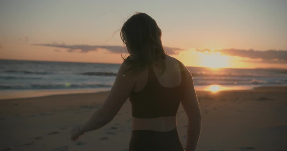 Young woman wearing yoga clothes walks on the sand beach at sunset next to the ocean
