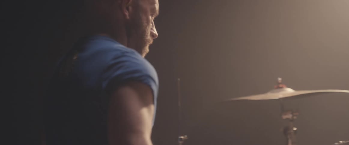 Bald male drummer with blue shirt and beard plays drums in spotlight