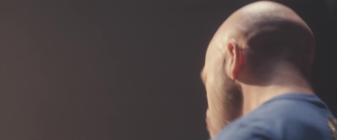 Bald male drummer with blue shirt and beard plays drums in spotlight