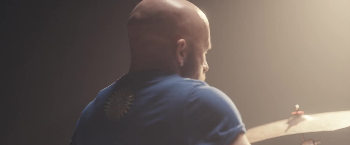 Bald male drummer with blue shirt and beard plays drums in spotlight