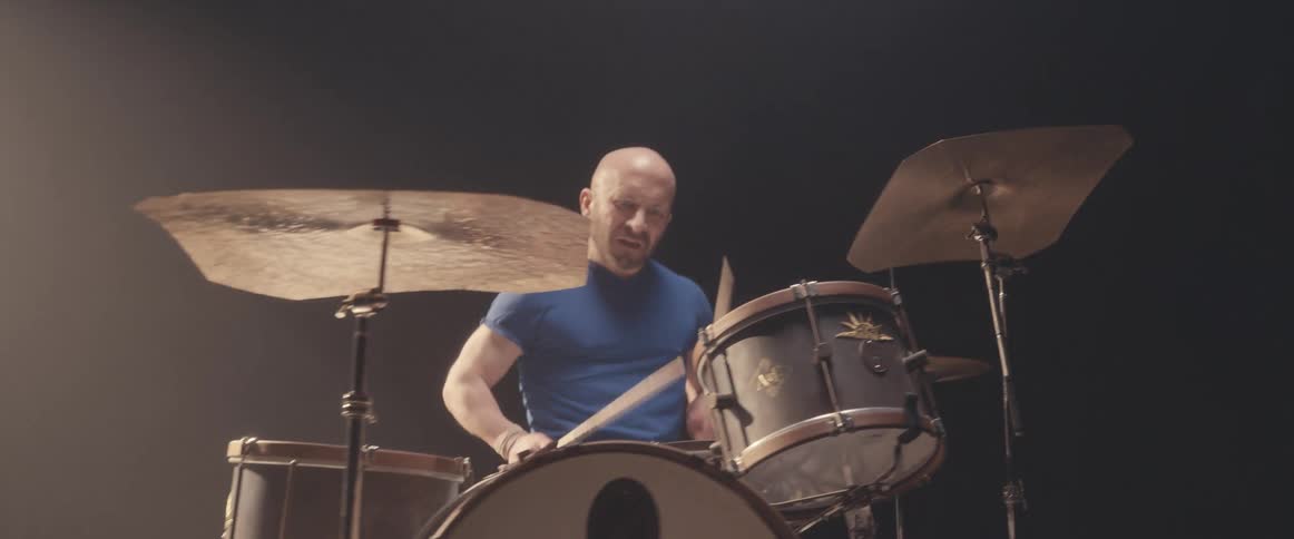 Bald male drummer with blue shirt and beard plays drums in spotlight