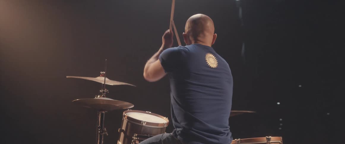 Bald male drummer with blue shirt and beard plays drums in spotlight