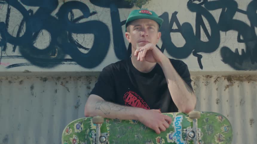 A man smoking a cigarette while sitting outdoors with a skateboard in his lap turns his head to look into the camera