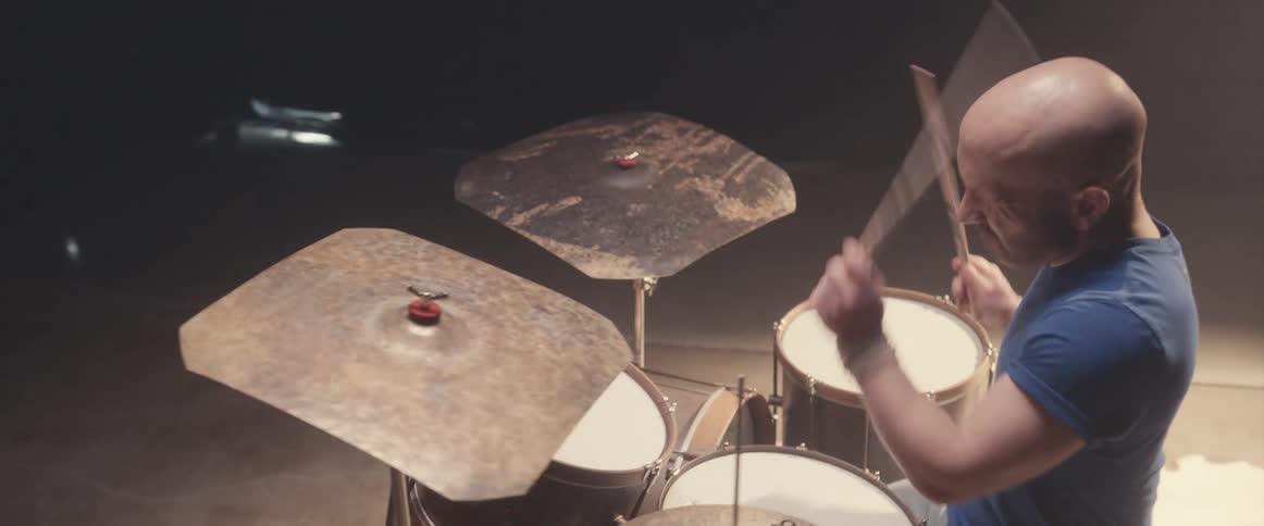 Bald male drummer with blue shirt jeans and beard plays drums in spotlight