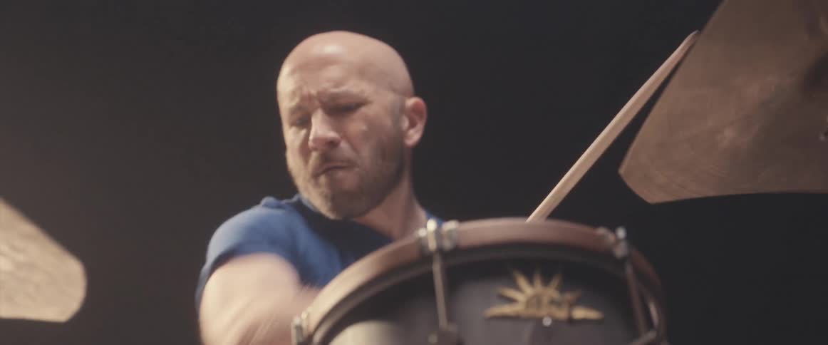 Bald male drummer with blue shirt and beard plays drums in spotlight