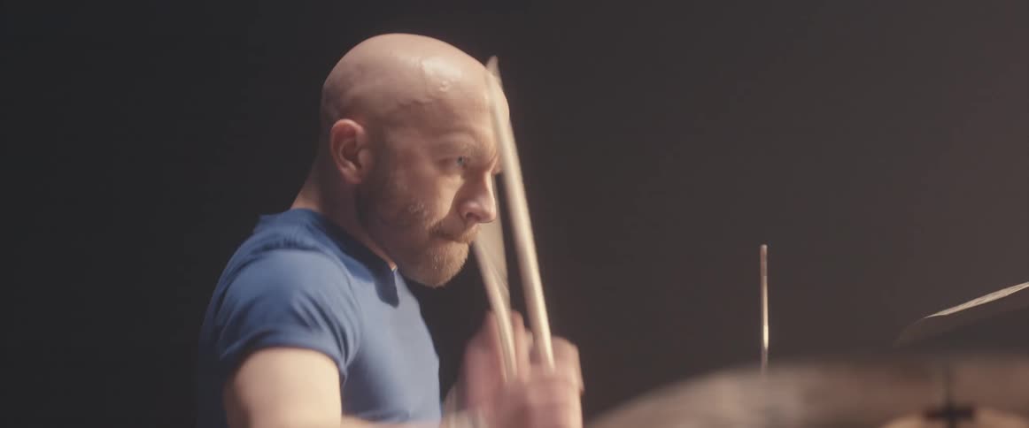 Bald male drummer with blue shirt and beard plays drums in spotlight