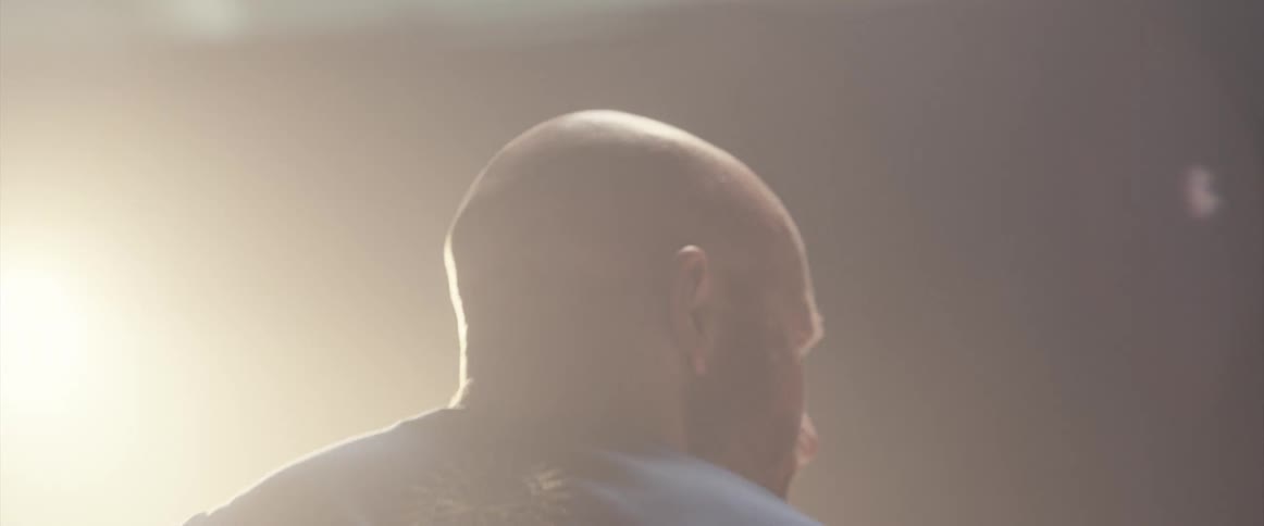 Bald muscular man energetically playing drums on a stage under a spotlight