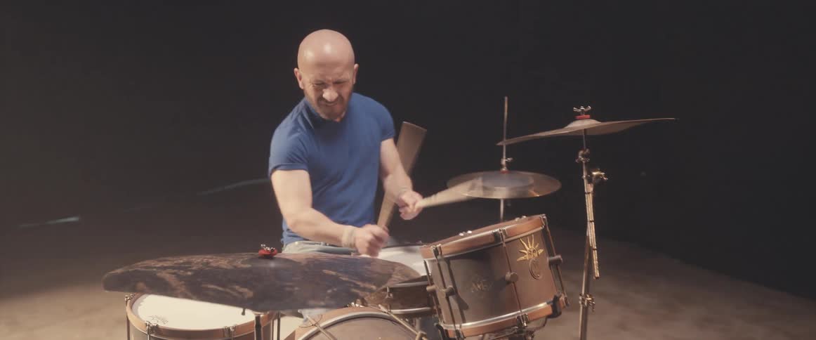 Bald male drummer with blue shirt and beard plays drums in spotlight