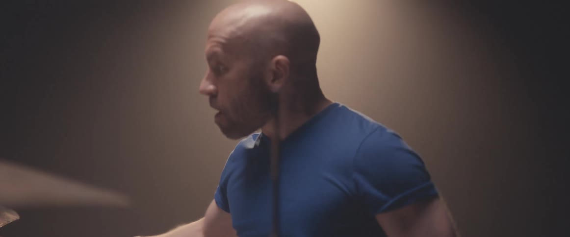 Bald male drummer with blue shirt and beard plays drums in spotlight