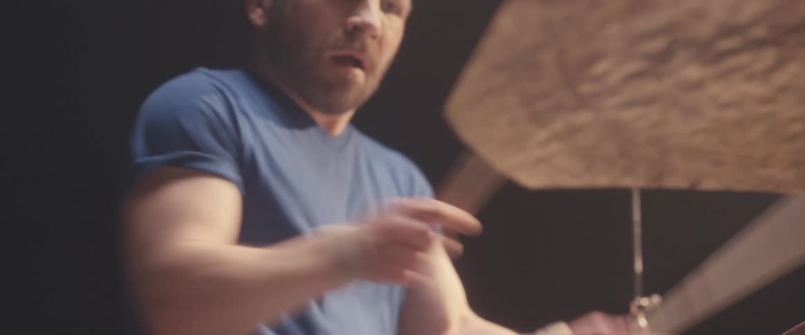 Bald male drummer with blue shirt and beard plays drums in spotlight