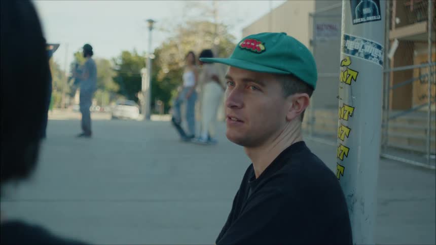 A man sitting leaning against a pole smoking a cigarette and laughing at the skate park