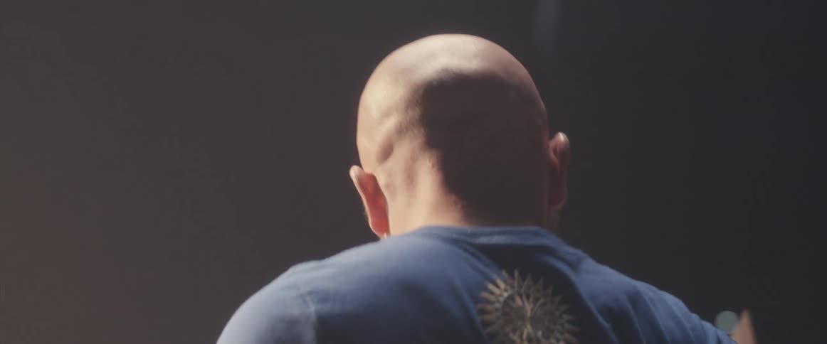Bald male drummer with blue shirt and beard plays drums in spotlight