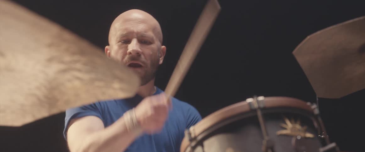 Bald male drummer with blue shirt and beard plays drums in spotlight