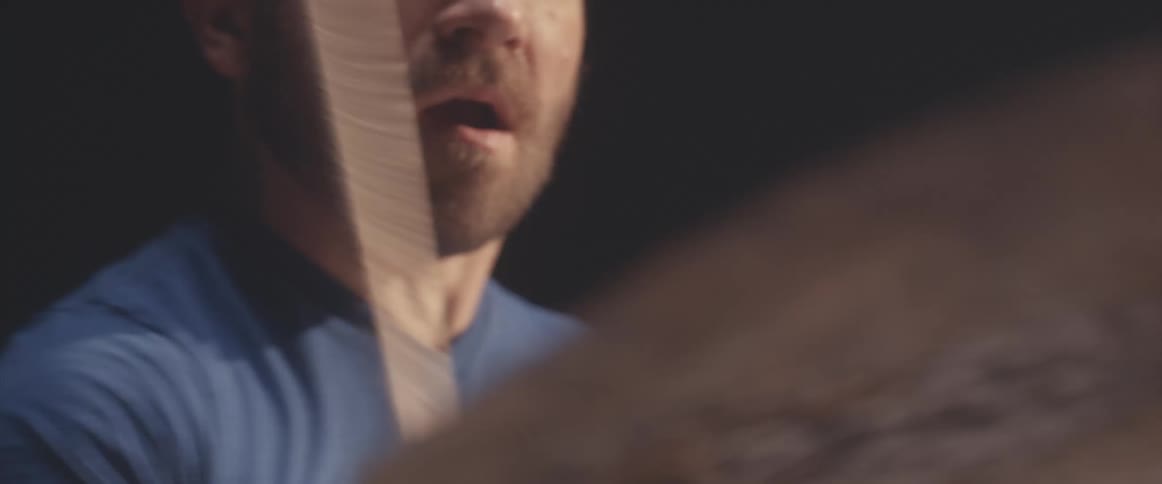 Bald male drummer with blue shirt and beard plays drums in spotlight