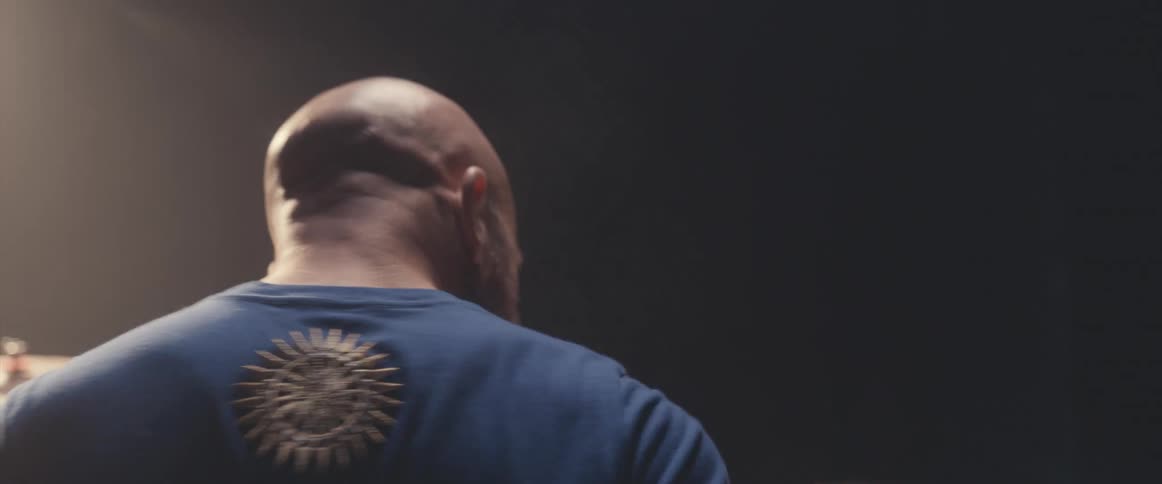 Bald male drummer with blue shirt and beard plays drums in spotlight