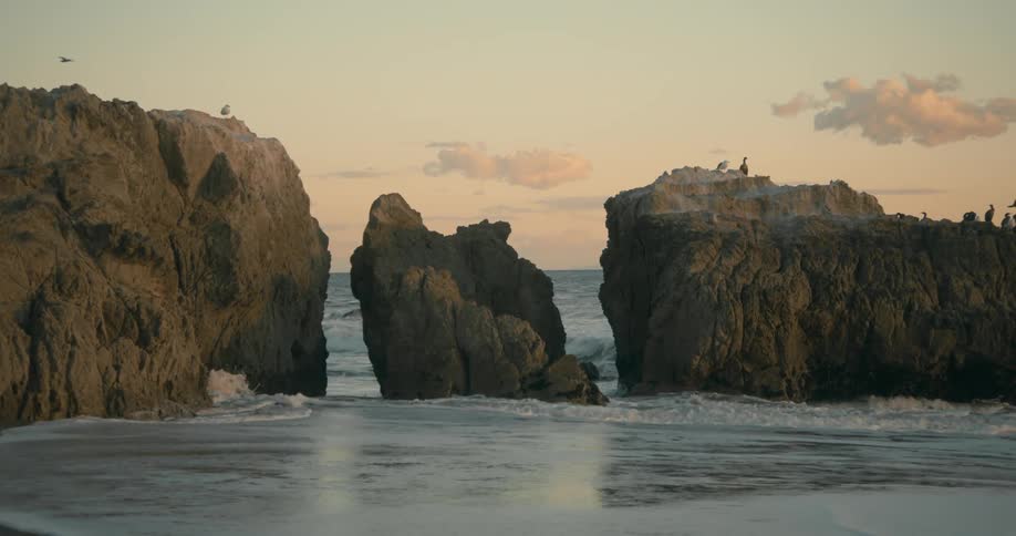 Water splashes against large rocks in the water as waves roll over the beach