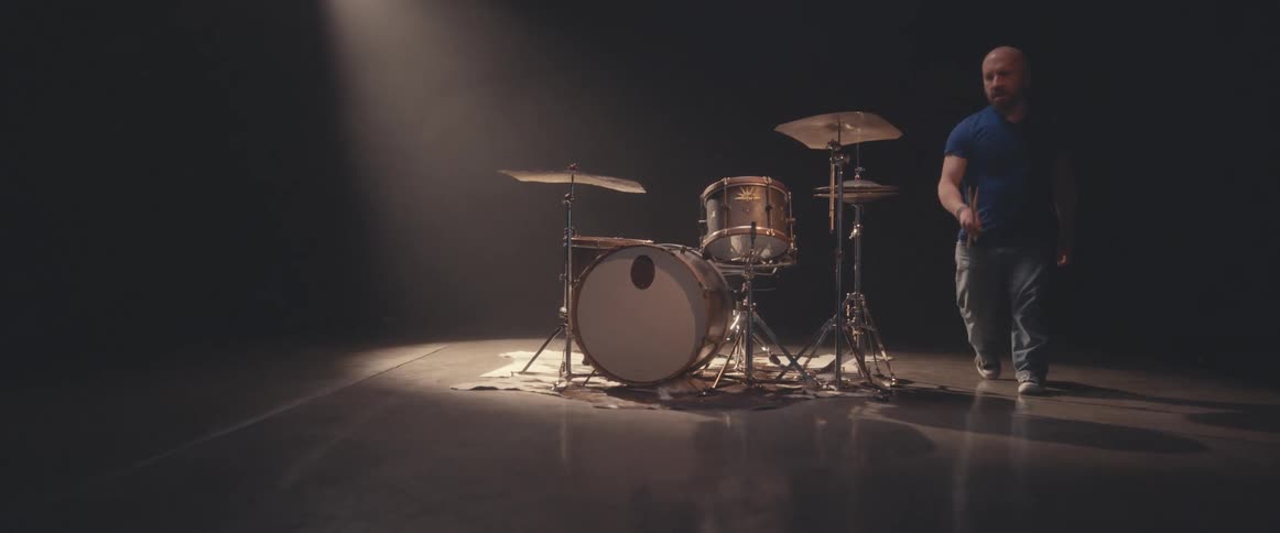 Bald male drummer with blue shirt and beard plays drums in spotlight
