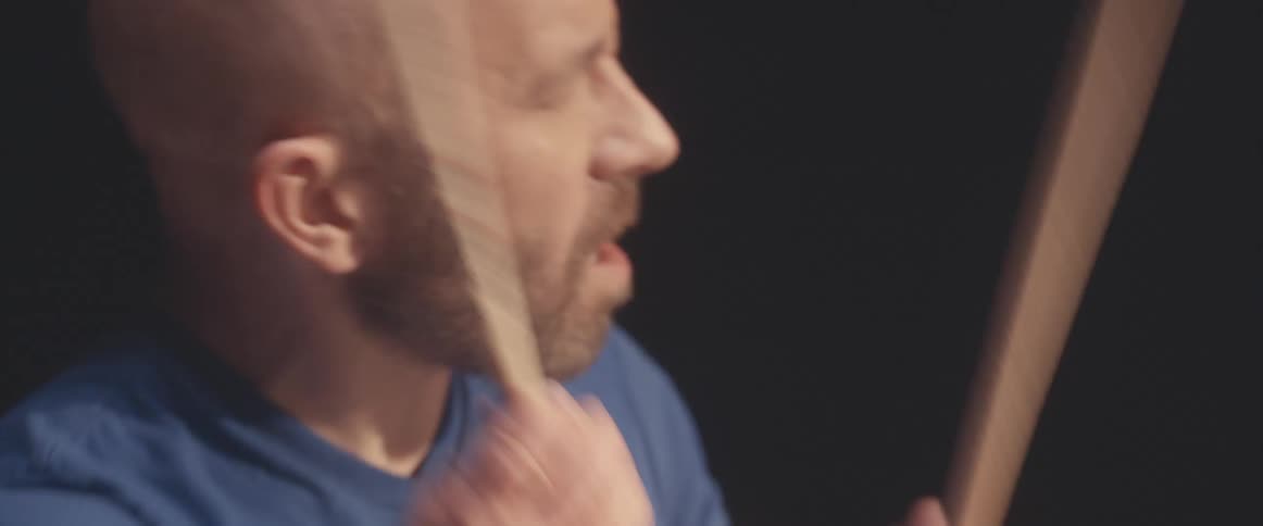 Bald male drummer with blue shirt and beard plays drums in spotlight