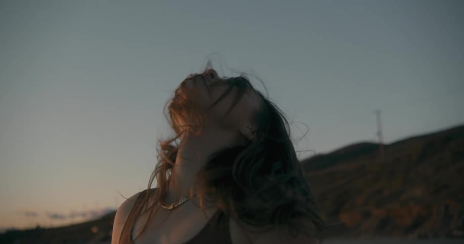 A young woman stretches her neck while standing on a beach at sunset