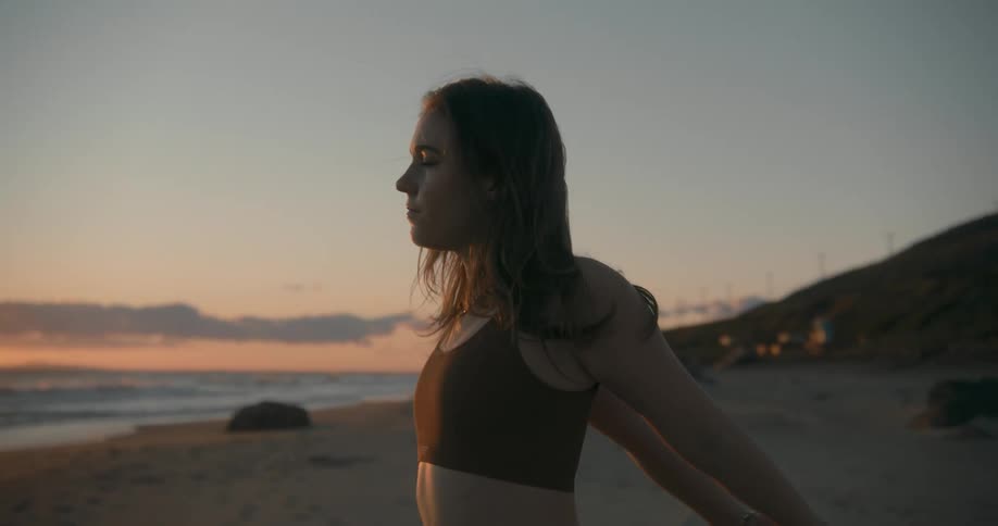 A young woman standing on the beach stretches her arms while looking out over the ocean at sunset