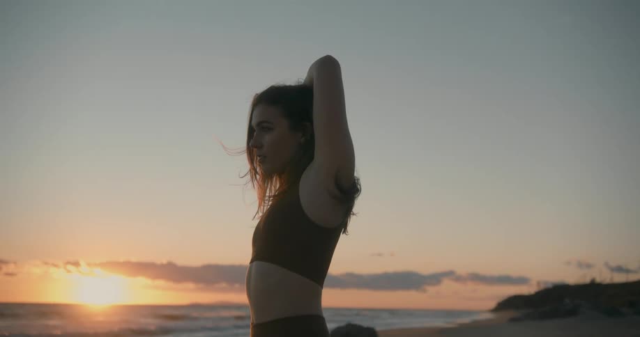 Young woman wearing yoga clothes stretching on the sand beach at sunset next to the ocean