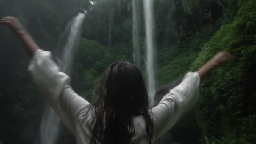 Young woman with wet hair stands in front of a waterfall on the mountains in the tropical jungle