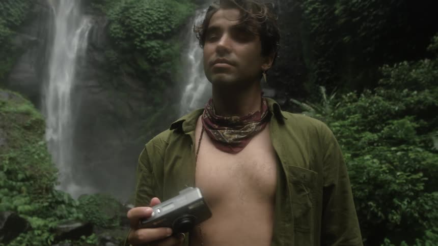 A man takes photos with his camera while standing in a rainforest in front of two waterfalls