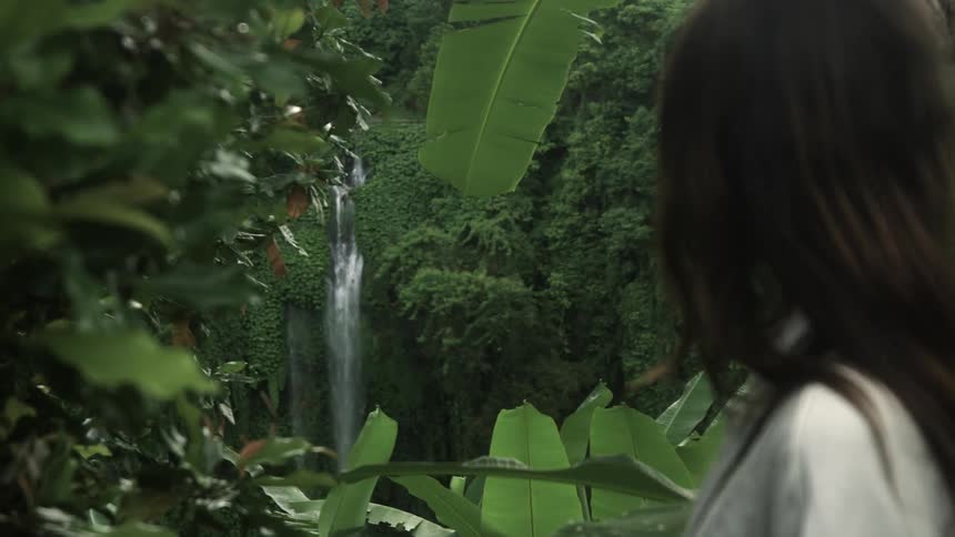 A woman pushes a large wet leaf down to get a clear view of the waterfall