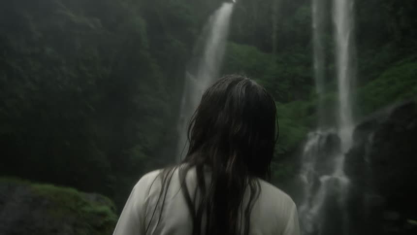 Woman with straight hair white blouse and green pants looks at waterfall cascading down cliffside in the jungle