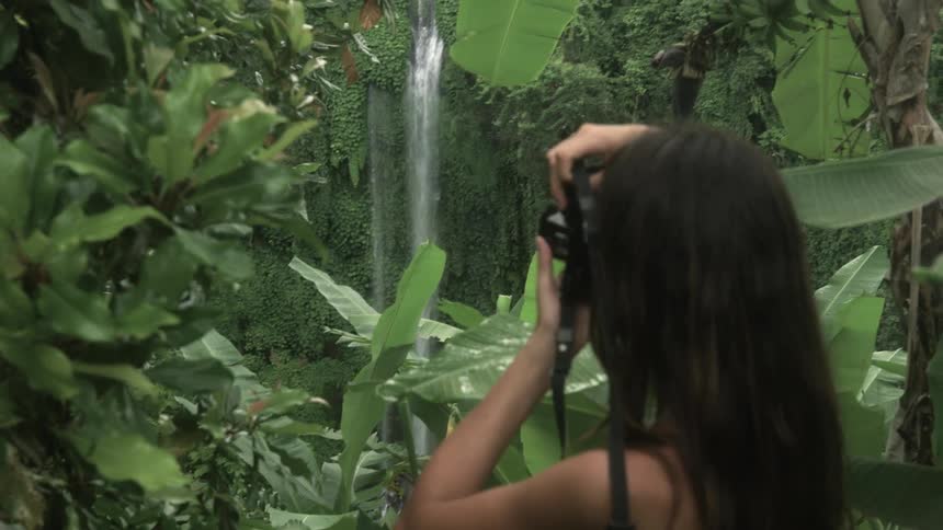 Young woman takes pictures of a waterfall on a tropical forest with a camera
