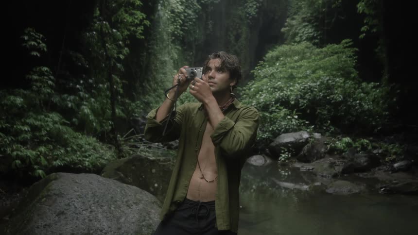 Man with green shirt earrings scarf and black pants photographs trees in the woods next to creek