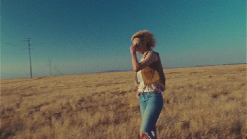 A woman in sunglasses walks through a vast field in the countryside during the day
